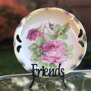 May include: A decorative porcelain plate with a hand-painted floral design featuring pink roses and green leaves. The plate has a scalloped edge with gold trim and two handle cutouts. The word "Friends" is displayed on a black stand.