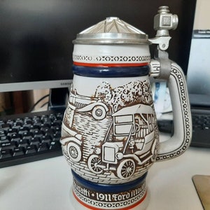 K&ouml;nnte beinhalten: Ein wei&szlig;er Keramik-Bierkrug mit einem blauen und roten Band um den oberen Rand. Der Krug zeigt eine detaillierte Illustration eines Oldtimers mit dem Text "1911 Ford Model T" auf der Unterseite.
