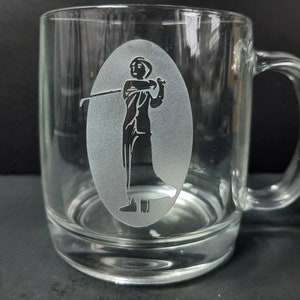 May include: A clear glass mug with a frosted design of a person holding a golf club. The mug has a handle on the right side.