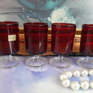 May include: Four red glass goblets with clear glass stems and bases. The goblets are arranged in a row on a light blue surface. A string of white pearls is in the foreground.