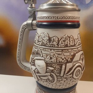 K&ouml;nnte beinhalten: Ein wei&szlig;er Keramik-Bierkrug mit einer braunen und wei&szlig;en Szene von Oldtimern, die an einer Backsteinmauer vorbeifahren. Der Krug hat einen silbernen Deckel und einen silbernen Henkel mit einem dekorativen Muster.