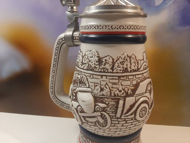 K&ouml;nnte beinhalten: Ein wei&szlig;er Keramik-Bierkrug mit einer braunen und wei&szlig;en Szene von Oldtimern, die an einer Backsteinmauer vorbeifahren. Der Krug hat einen silbernen Deckel und einen silbernen Henkel mit einem dekorativen Muster.