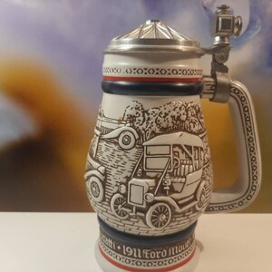 K&ouml;nnte beinhalten: Ein wei&szlig;er Keramik-Bierkrug mit Deckel und Henkel. Der Krug zeigt ein Reliefdesign von Oldtimern und den Text "1911 Ford Model T".