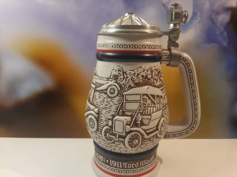 K&ouml;nnte beinhalten: Ein wei&szlig;er Keramik-Bierkrug mit Deckel und Henkel. Der Krug zeigt ein Reliefdesign von Oldtimern und den Text "1911 Ford Model T".