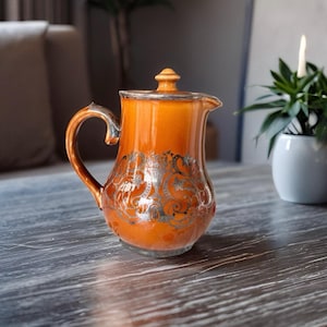 May include: An orange ceramic pitcher with a silver rim, handle, and decorative floral pattern. The pitcher has a lid and a spout for pouring. The design is vintage and elegant, suitable for serving beverages.
