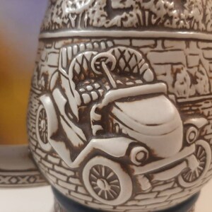K&ouml;nnte beinhalten: Eine wei&szlig;e Keramik-Bierkr&uuml;ge mit einem detaillierten Reliefdesign eines Oldtimers. Das Auto ist von einer Backsteinmauer und Laub umgeben.