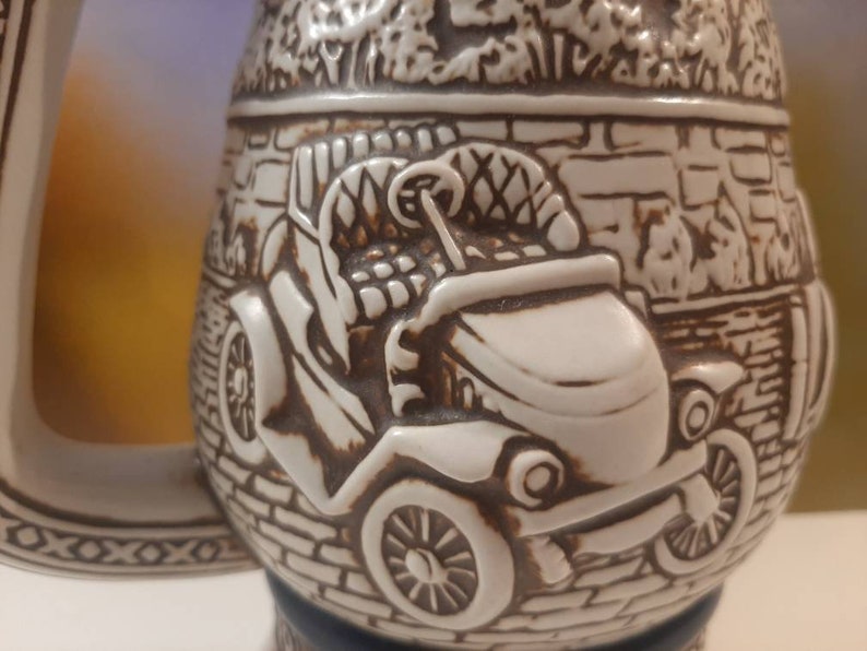 K&ouml;nnte beinhalten: Eine wei&szlig;e Keramik-Bierkr&uuml;ge mit einem detaillierten Reliefdesign eines Oldtimers. Das Auto ist von einer Backsteinmauer und Laub umgeben.
