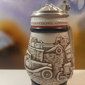 K&ouml;nnte beinhalten: Ein wei&szlig;er Bierkrug aus Keramik mit einem Zinnverschluss. Der Krug zeigt eine detaillierte Illustration von Oldtimern und einer Backsteinmauer. Der Deckel hat ein rotes und blaues Band mit einem sich wiederholenden Muster aus X.