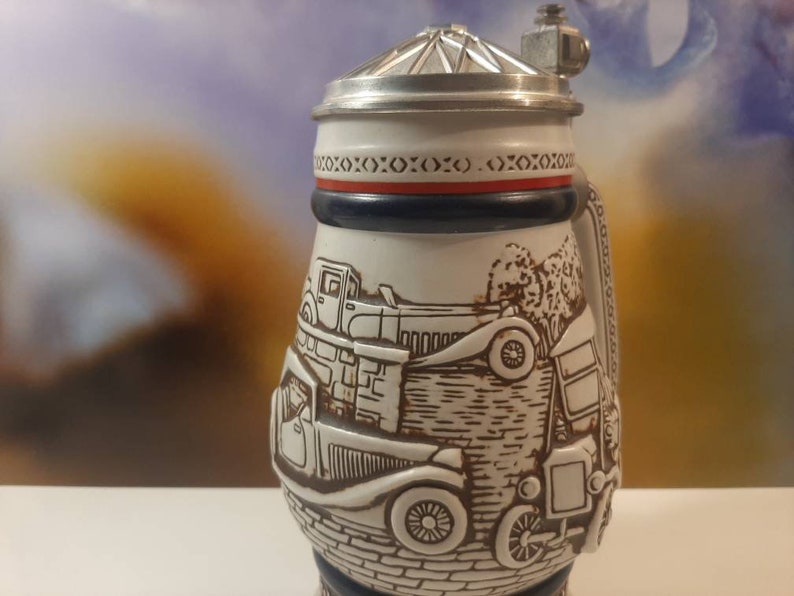 K&ouml;nnte beinhalten: Ein wei&szlig;er Bierkrug aus Keramik mit einem Zinnverschluss. Der Krug zeigt eine detaillierte Illustration von Oldtimern und einer Backsteinmauer. Der Deckel hat ein rotes und blaues Band mit einem sich wiederholenden Muster aus X.