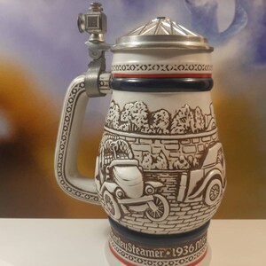 K&ouml;nnte beinhalten: Ein wei&szlig;er Keramik-Bierkrug mit einer braunen und wei&szlig;en Szene von Oldtimern und B&auml;umen. Der Krug hat einen Zinn-Deckel und einen Metallgriff mit einer kleinen Metallfigur oben. Der Krug tr&auml;gt den Text "Steamers 1936 Mug" auf der Unterseite.