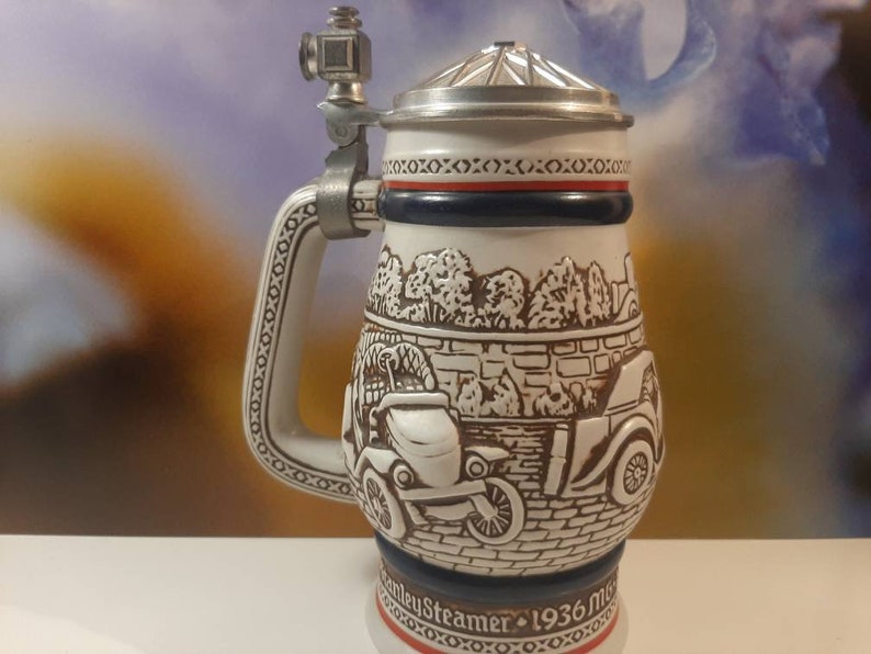 K&ouml;nnte beinhalten: Ein wei&szlig;er Keramik-Bierkrug mit einer braunen und wei&szlig;en Szene von Oldtimern und B&auml;umen. Der Krug hat einen Zinn-Deckel und einen Metallgriff mit einer kleinen Metallfigur oben. Der Krug tr&auml;gt den Text "Steamers 1936 Mug" auf der Unterseite.