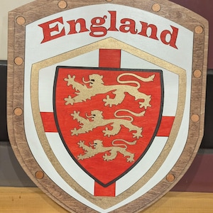 May include: Wooden shield featuring the word "England" in red. The shield has a red and white design with three gold lions. The shield is framed with a brown border and decorative rivets.