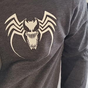 May include: A dark gray hooded sweatshirt with a white Venom graphic on the front. The graphic features a white spider with a white Venom face.