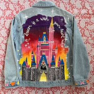 May include: A denim jacket with a painted design of a castle with a pink and purple sky. The text "A dream is a wish your heart makes" is written on the jacket.