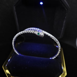 Könnte beinhalten: Ein silbernes Schlangenarmband mit einem grünen Edelsteinauge. Das Armband befindet sich in einer blauen Samtbox.