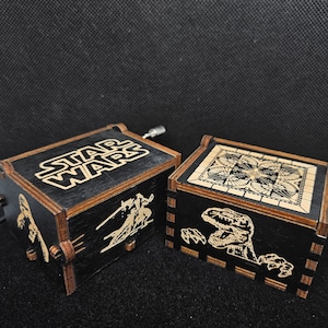 May include: Two wooden music boxes with intricate designs. One box features the Star Wars logo and characters, while the other has a dinosaur design.
