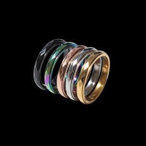 May include: Seven faceted rings in a range of colours, including black, iridescent, rose gold, silver, and gold. The rings are arranged in a curved line against a black background, highlighting their geometric design.