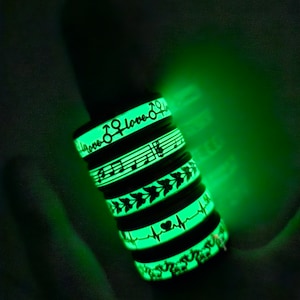 Könnte beinhalten: Ein Stapel von fünf im Dunkeln leuchtenden Armbändern mit verschiedenen Designs. Die Armbänder zeigen Symbole wie Noten, Herzen und die Wörter "love" und "peace".