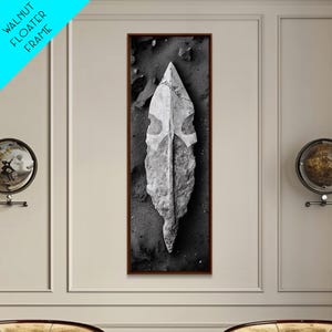 May include: Black and white framed art print featuring a detailed image of a weathered, stone arrowhead. The arrowhead is set against a dark, textured background, with a walnut floater frame. The artwork is displayed on a neutral wall.