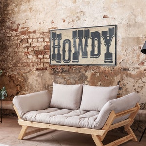 May include: A light beige futon sofa with wooden legs and two pillows. The sofa is in front of a brick wall with a sign that reads "HOWDY" in black letters.