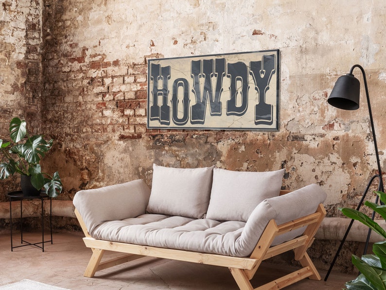 May include: A light beige futon sofa with wooden legs and two pillows. The sofa is in front of a brick wall with a sign that reads "HOWDY" in black letters.