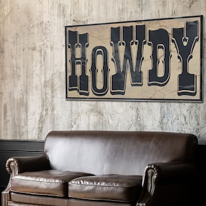 May include: A rustic wooden sign with the word "HOWDY" in black lettering on a beige background. The sign is hanging on a textured gray wall above a brown leather couch.