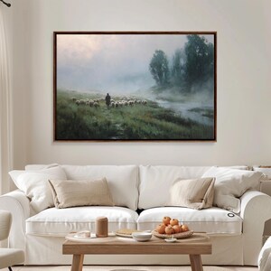 May include: A framed landscape painting depicts a shepherd and flock of sheep in a misty, green field. A river runs along the right side, with trees in the background. The artwork has a dark brown frame and is displayed above a white sofa.