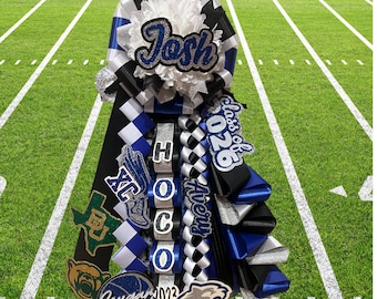 May include: A blue, black, and white graduation lei with the name "Josh" and the text "Class of 2026" on it. The lei also has a white flower, a silver ribbon, and a black and white checkered ribbon. The lei is on a green football field.