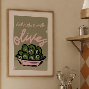 Op de afbeelding: Een ingelijste wandkunstprint met een saliegroene achtergrond en een kom met groene olijven. De tekst Let's start with olives staat in roze schrift. De illustratie bevat kleine roze steraccenten en een roze kom, gevat in een lichthouten lijst.