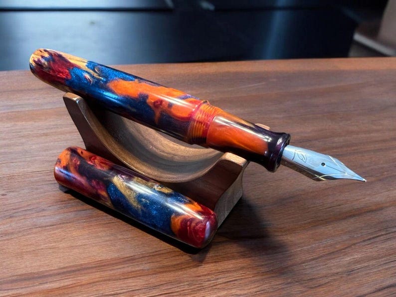 May include: Close-up view of two handcrafted resin fountain pens displayed on a dark brown wooden stand. The pens feature vibrant, multicolored resin bodies with swirling patterns of deep blues, oranges, and reds. The nibs are silver-toned.  The pens are showcased as high-end writing instruments, highlighting their unique design and craftsmanship.