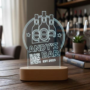 Może przedstawiać: Podświetlany szyld barowy z napisem "ANDY'S BAR EST 2023" i kodem QR. Znak ma okrągły kształt z trzema ilustracjami butelek i akcentami gwiazd. Spoczywa na drewnianej podstawie i emituje delikatną poświatę.