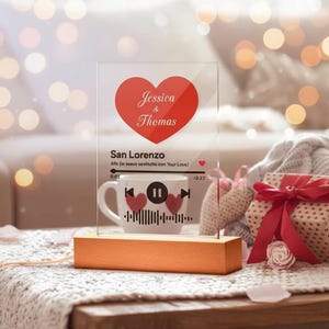 May include: A clear acrylic plaque on a wooden base, with a red heart bearing the names "Jessica & Thomas." The text "San Lorenzo" and a music waveform design are displayed. A white mug with a music-themed design is also visible.