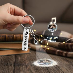 May include: A silver keychain with a rectangular tag displaying a music code and a small charm. The keychain is attached to a small chain and a round key ring. The image also shows a projected image of a couple and the words "CUSTOM Music".