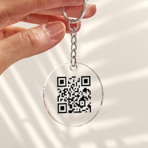 May include: Transparent, circular keyring featuring a black QR code. The keyring includes a silver-coloured chain and ring for attachment. The QR code is centrally positioned on the keyring.