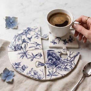 May include: A white ceramic puzzle-shaped coaster set with a blue floral design, holding a white coffee cup filled with dark coffee. The coaster set is composed of four pieces. A spoon and blue flower are also visible.
