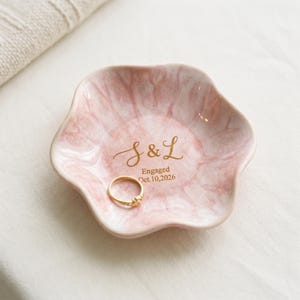May include: A flower-shaped ceramic ring dish with a pink and white marbled finish. The center features gold lettering that reads S & L, Engaged, Oct. 10, 2026. A thin gold ring rests inside the dish, which has wavy, scalloped edges and a smooth surface.