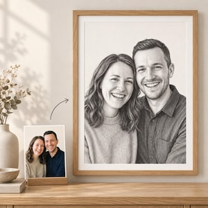 May include: A framed black and white pencil sketch portrait of a smiling couple, displayed on a wooden surface next to a smaller original colour photograph of the same two people. The artwork is encased in a light-toned wooden frame.