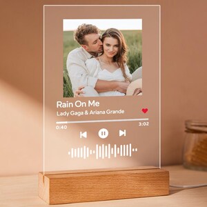 May include: A clear acrylic plaque with a wooden base, featuring a photo of a couple. The plaque displays the song title "Rain On Me" by Lady Gaga & Ariana Grande, along with a play bar and music waveform. The base is rectangular and made of light-colored wood.