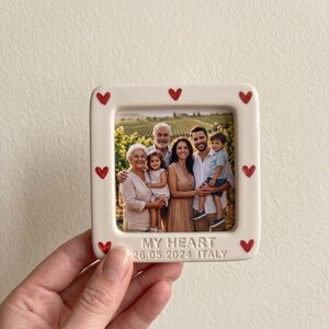 Pode incluir: Uma moldura quadrada de cer&acirc;mica em tom esbranqui&ccedil;ado com uma fotografia de uma fam&iacute;lia de cinco pessoas num vinhedo. A moldura &eacute; decorada com pequenos cora&ccedil;&otilde;es pintados de vermelho e inclui o texto em relevo MY HEART 26.05.2024 ITALY.