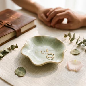 May include: A pale green ceramic ring dish with a wavy, flower-like edge. The center features gold printed text that reads e + b and the date 2026.11.12. A thin, twisted gold ring rests inside the dish, which has a smooth, glossy finish.