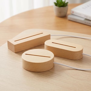 May include: Three wooden LED light bases in various shapes: rectangular, oval, and circular. Each base features a slot for a light panel and a white cable. The bases are crafted from light-coloured wood and are placed on a wooden surface.