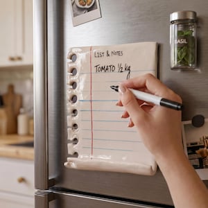 May include: A ceramic notepad-shaped magnet with the text "LIST & NOTES" and "TOMATO 1/2 kg" written on it. A hand is holding a black marker, writing on the notepad. A jar of basil and a photo are also attached to the refrigerator.