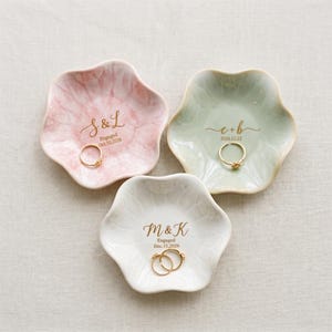 May include: Three flower-shaped ceramic ring dishes in pastel pink, sage green, and white. Each dish features gold-colored custom text including initials, the word Engaged, and a date. A gold ring rests in the center of each individual dish.