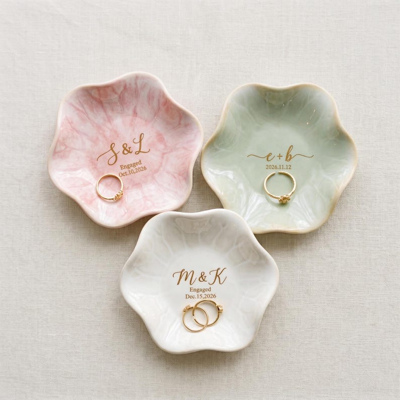 May include: Three flower-shaped ceramic ring dishes in pastel pink, sage green, and white. Each dish features gold-colored custom text including initials, the word Engaged, and a date. A gold ring rests in the center of each individual dish.