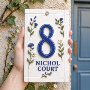 May include: A rectangular white ceramic house sign featuring a large embossed blue number 8 in the centre. Below the number, the text NICHOL COURT is printed in blue. The sign is decorated with raised blue and white floral motifs and green stems.