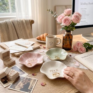 May include: Three scalloped ceramic trinket dishes in pink, light green, and white, each personalized with gold lettering featuring initials and dates. The dishes are arranged on a wooden surface alongside a beige ring box, a mug with the text I Said Yes, and a glass vase containing pink roses.