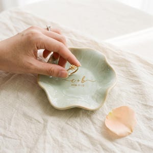 May include: A pale green, flower-shaped ceramic ring dish featuring the gold printed text e + b and the date 2026.11.12. The dish has a smooth, glossy finish with wavy, scalloped edges and a shallow, rounded basin for holding small jewelry items.