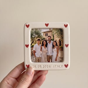 Pode incluir: Uma moldura quadrada de cer&acirc;mica em tom off-white com acabamento brilhante e cora&ccedil;&otilde;es vermelhos pintados &agrave; m&atilde;o na borda. A moldura exibe uma fotografia de quatro pessoas e inclui o texto em relevo 26.05.2024 ITALY na borda inferior.