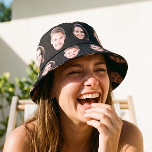Może przedstawiać: Czarny kapelusz typu bucket hat z powtarzającym się wzorem przedstawiającym różne ludzkie twarze nadrukowane na tkaninie. Kapelusz ma szerokie, opadające rondo i zaokrągloną główkę, wykonany z ciemnego materiału tekstylnego z wieloma portretami.