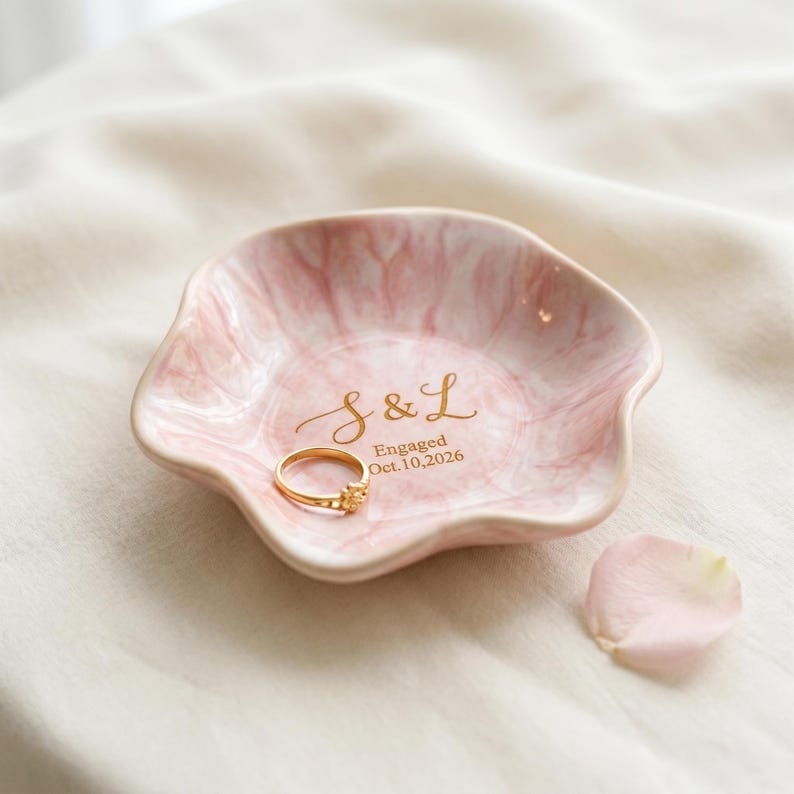 May include: A scalloped, pink and white marbled ceramic ring dish featuring gold lettering that reads S & L Engaged Oct. 10, 2026. A thin gold ring rests inside the dish, and a single pale pink flower petal is positioned nearby on the fabric surface.